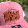 A heather red small trucker hat with a beige mesh back & a leather patch engraved with 'bless your heart'