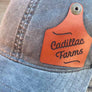 Blue cap with a brown cow tag shaped leather patch featuring 'Cadillac Farms' on a wooden background