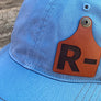 Blue cap with a brown cow tag shaped leather patch featuring the letter 'R' on a wooden surface