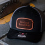 A black & gray trucker hat with a 'your logo design here' leather patch, sitting on a saw bench in a workshop