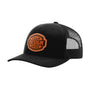 A black trucker-style cap with a curved brim, black mesh back, and a customizable brown patch on the front that reads 'YOUR DESIGN HERE'.