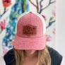 A woman wears a heather red small trucker hat with a beige mesh back & a leather patch engraved with 'bless your heart' 