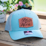 Light blue trucker cap with 'Bless your Heart' leather patch on a wooden surface with flowers in the background