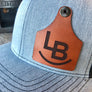 Blue trucker hat with a brown cattle tag shaped leather patch featuring 'LB' on a wooden surface.