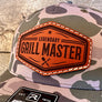 a tan & green camo hat with a brown, hexagon leather patch that reads "grill master"