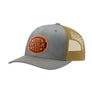 A grey trucker-style cap with a gold mesh back and a brown 'your design here' leather patch on the front panel, shown on a plain white background.