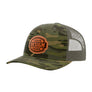 A camouflage trucker hat with a green and brown camo pattern, featuring a custom brown circular patch on the front that reads 'YOUR DESIGN HERE'. The back of the cap has a green mesh design.