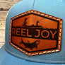 A light blue trucker hat with a hexagon shaped leather patch, engraved with 'reel joy' and a fish & boat.