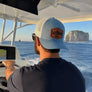 Man wearing a blue leather patch hat with the leather patch reading 'reel joy', drives a boat with a scenic background of cliffs and water.
