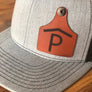 Gray cap with a brown cattle tag shaped leather patch featuring a black 'P' on a wooden surface